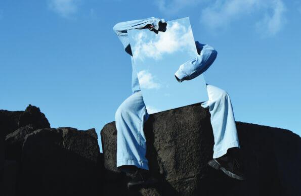 An artist in a blue suit sits on a rock with a blue sky behind