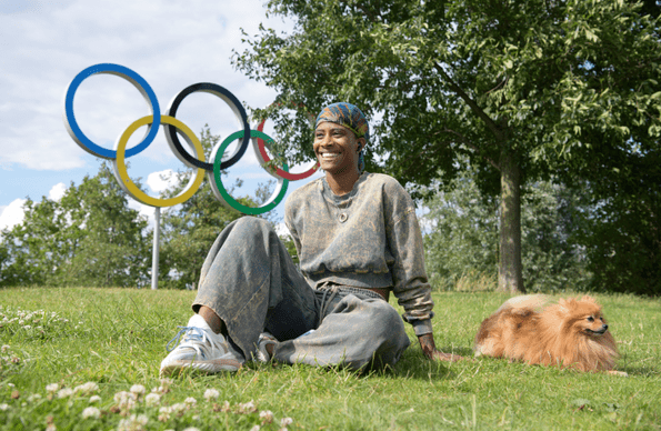 A man sits with his dog on the grass, with the Olympic rings in the background