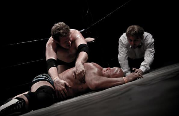 A man lies prone on the cavas of a wrestling ring with his opponent and the referee above him